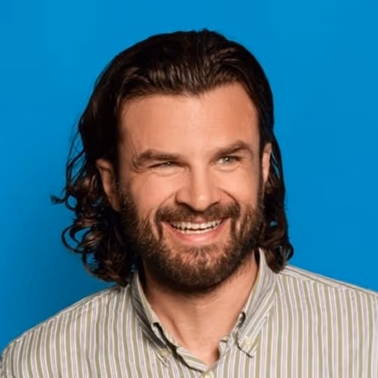 Smiling man with medium-length curly dark hair and beard wearing a striped button-up shirt against a blue background.