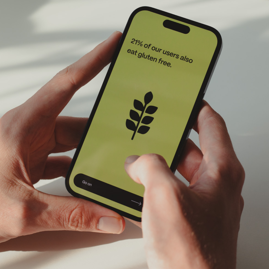 Hand holding a smartphone displaying the text '21% of our users also eat gluten free' with a black leaf icon on a light green background.
