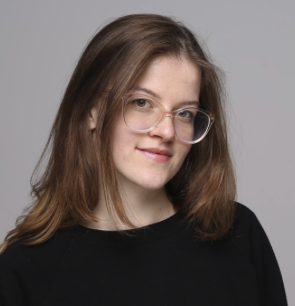 Smiling woman with light brown hair wearing glasses and a black top against a gray background.