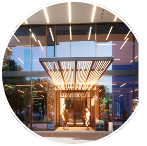 People standing under a modern illuminated canopy entrance of a glass building during dusk.