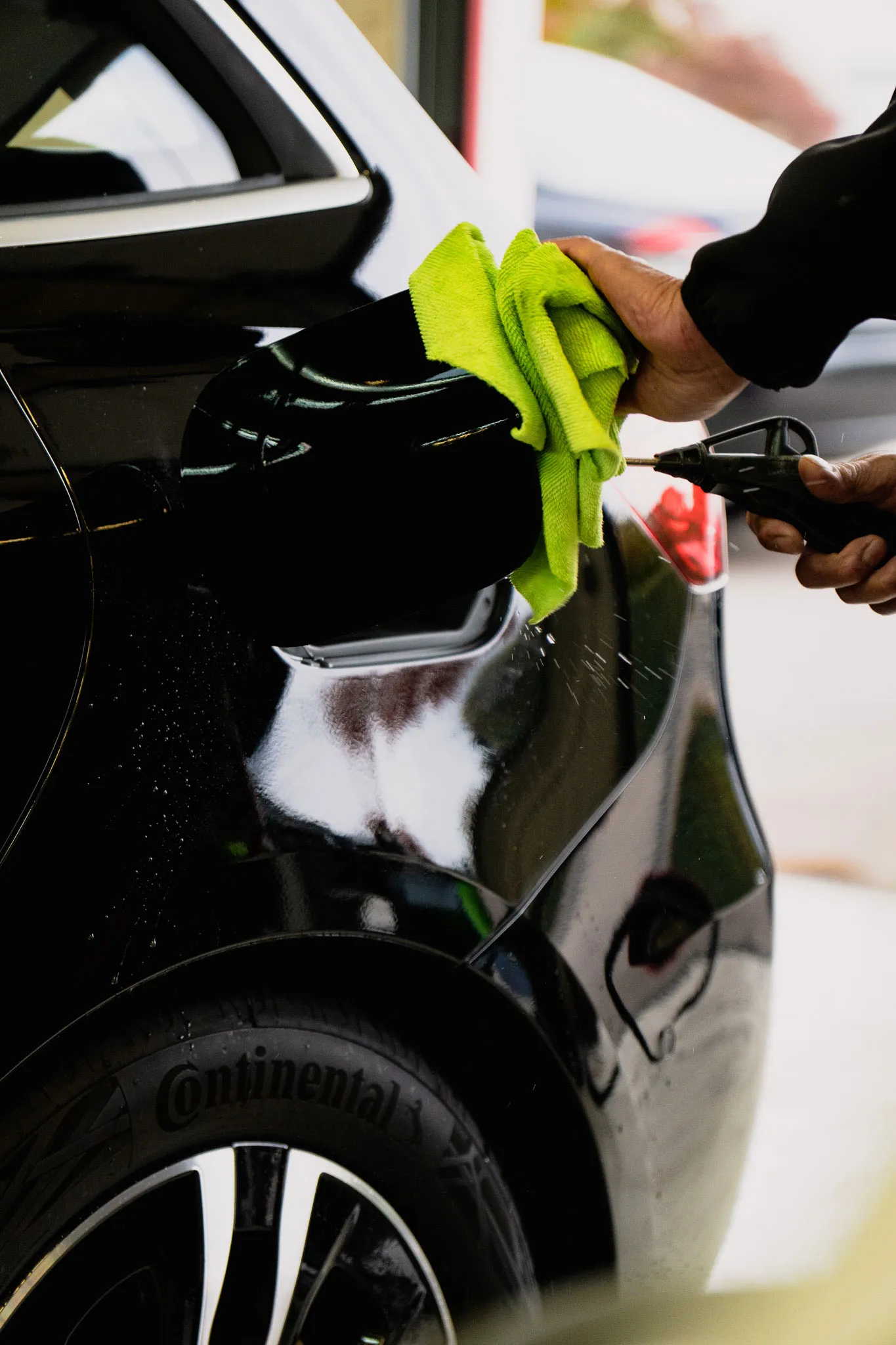 Person, der mit einem grünen Tuch einen schwarzen Autospiegel reinigt und mit einem Wasserstrahl abspritzt.