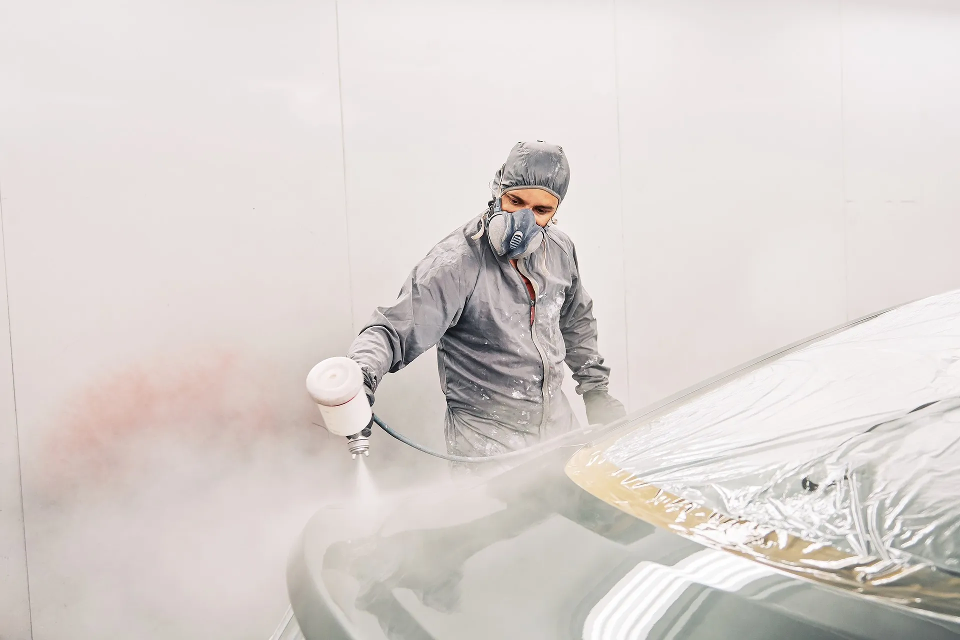 Mann in Schutzanzug und Maske lackiert ein Auto mit Spritzpistole in einer Lackierkabine.