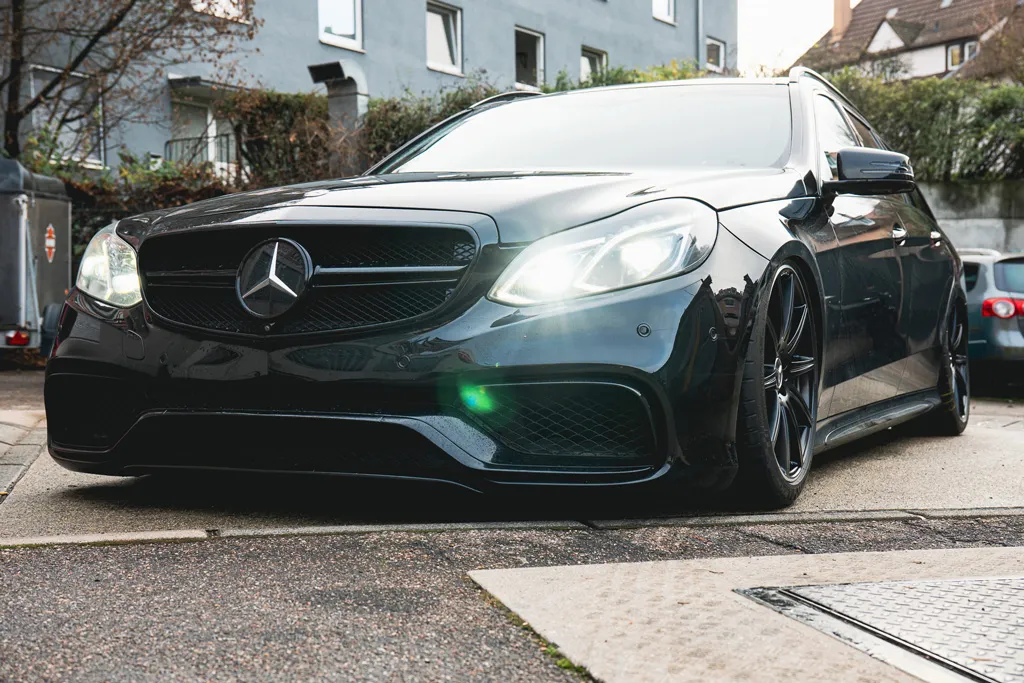 Schwarzer Mercedes mit eingeschaltetem Scheinwerfer, niedrigem Fahrwerk und schwarzen Felgen, geparkt auf einem Bürgersteig in Wohngegend.