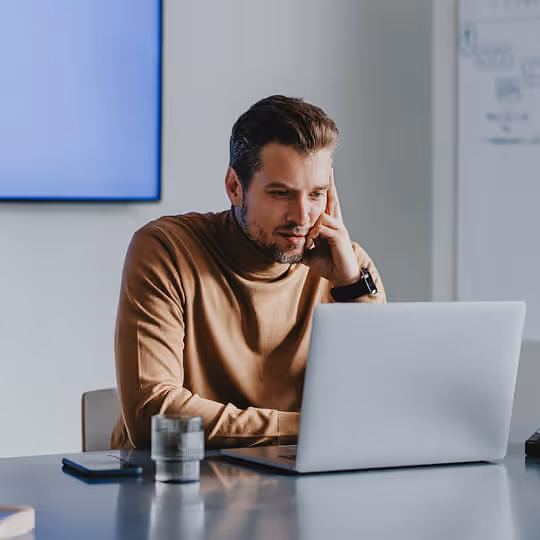Homem sentado em mesa moderna trabalhando concentrado em um laptop prateado, com celular e copo de vidro na mesa.