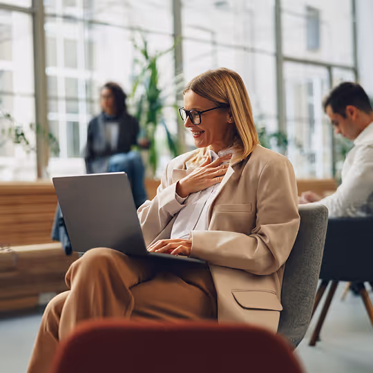 Mulher sorridente usando laptop em ambiente de trabalho moderno com outras pessoas ao fundo.