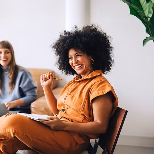 Mulher sorridente com cabelo afro usando roupa laranja, segurando uma caneta e um caderno, sentada em uma cadeira, com outra pessoa ao fundo.