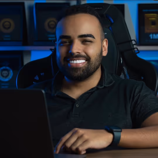 Homem sorridente sentado em cadeira gamer, em frente a um laptop, com placas de reconhecimento ao fundo.
