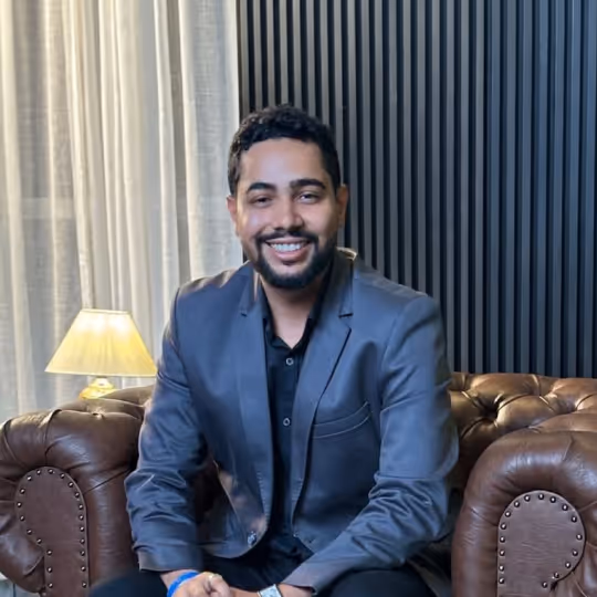 Homem sorridente vestido com terno cinza sentado em sofá de couro marrom.