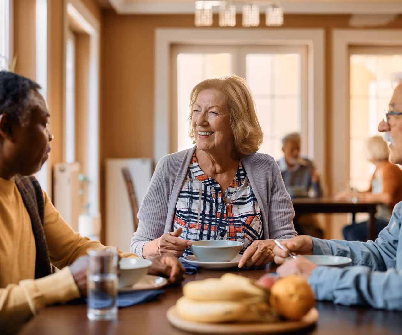 Three elderly people sitting at a table, smiling and holding bowls, enjoying a meal together in a warm, well-lit room.
