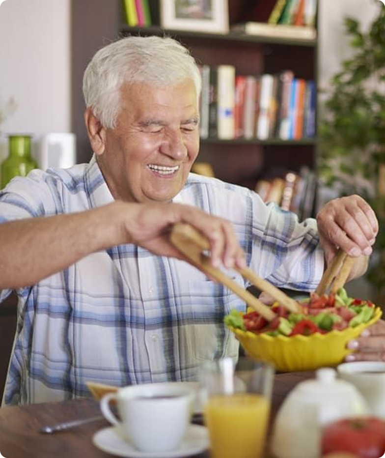 Smiling elderly man in a plaid shirt serving salad from a yellow bowl at a dining table with a cup of coffee and glass of orange juice.