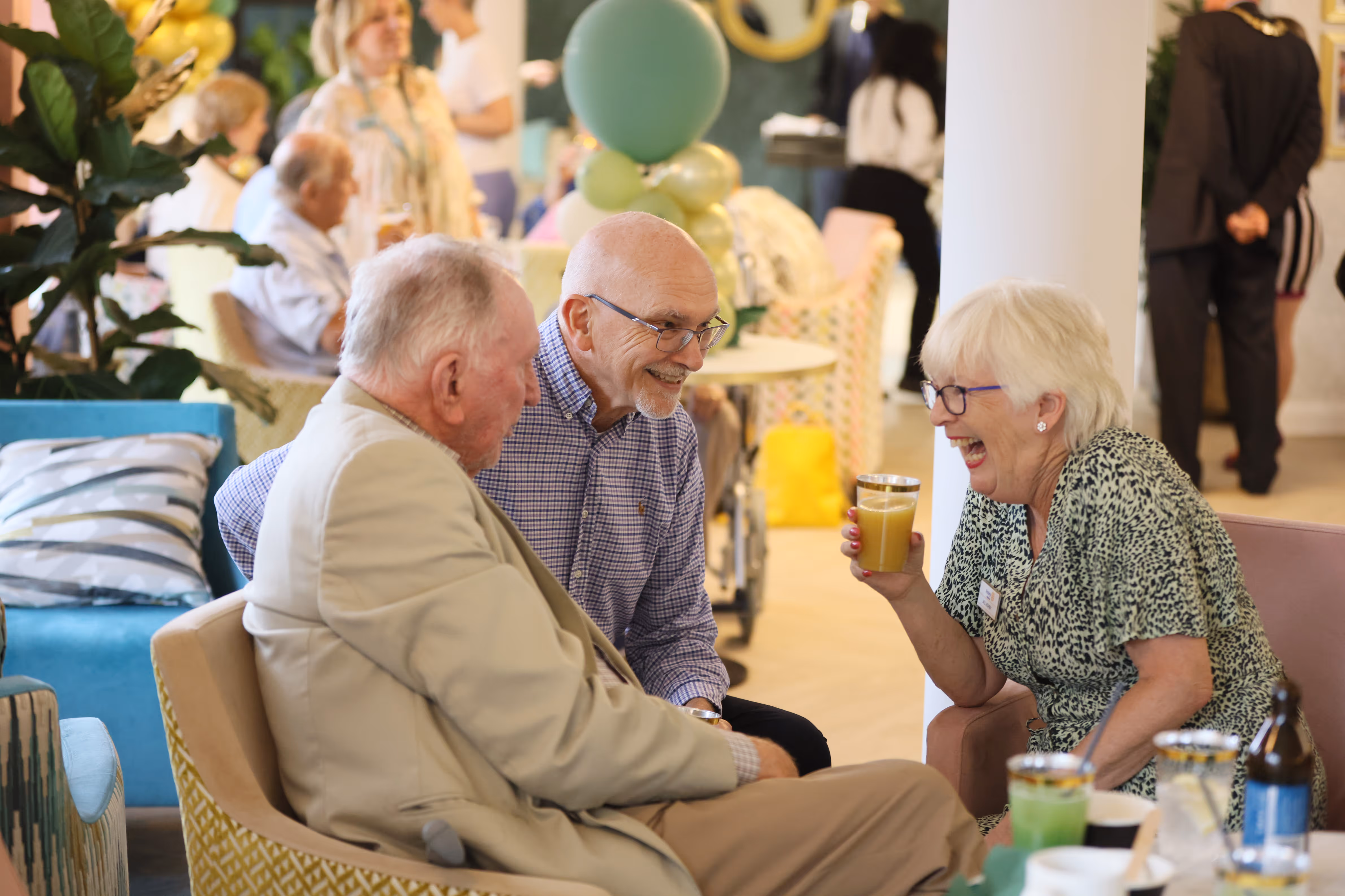 Three elderly people sitting and smiling together in a social setting, one woman holding a glass of orange juice, surrounded by decorations and other people in the background.