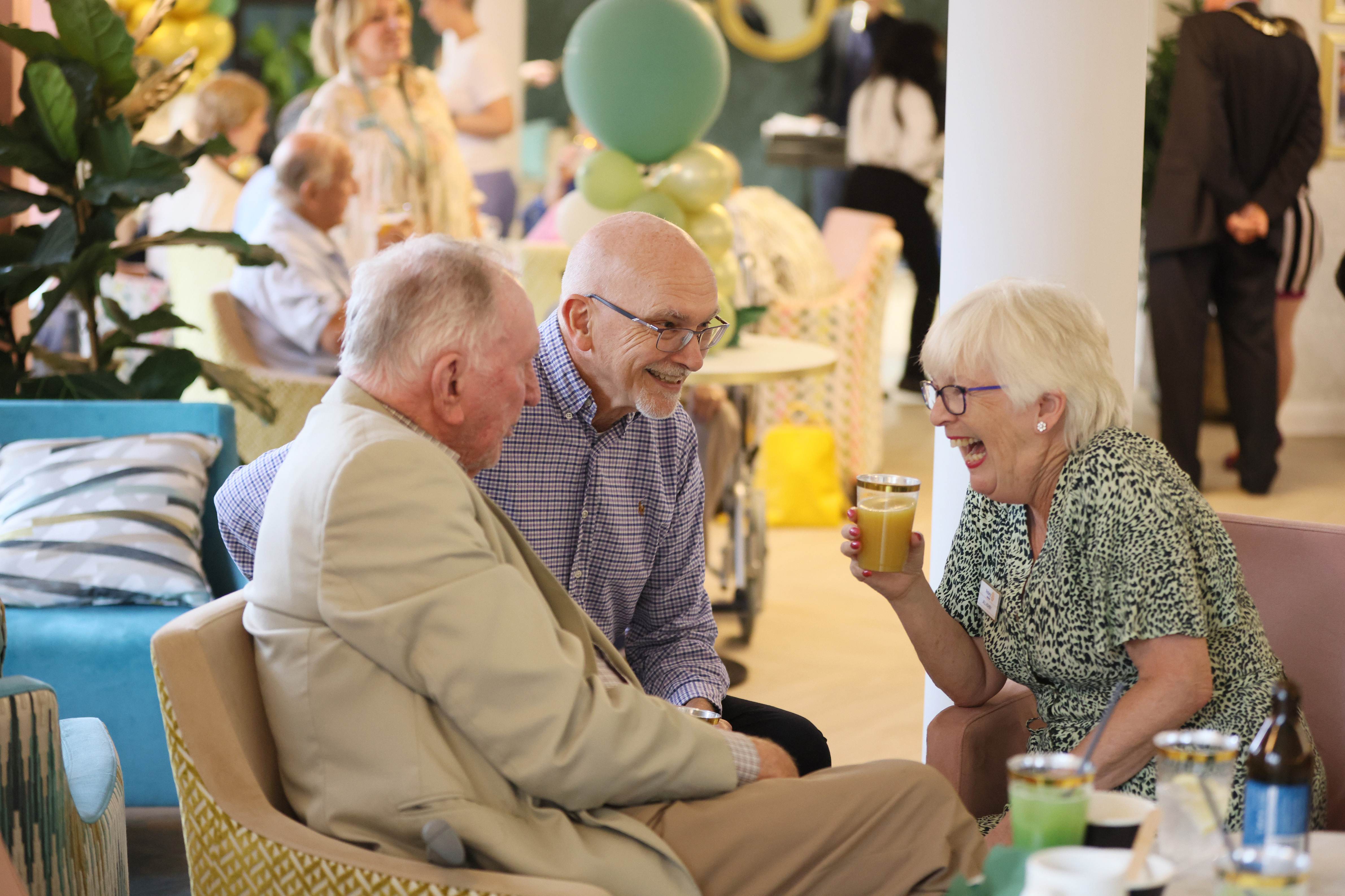 Three elderly people sitting and smiling together in a social setting, one woman holding a glass of orange juice, surrounded by decorations and other people in the background.