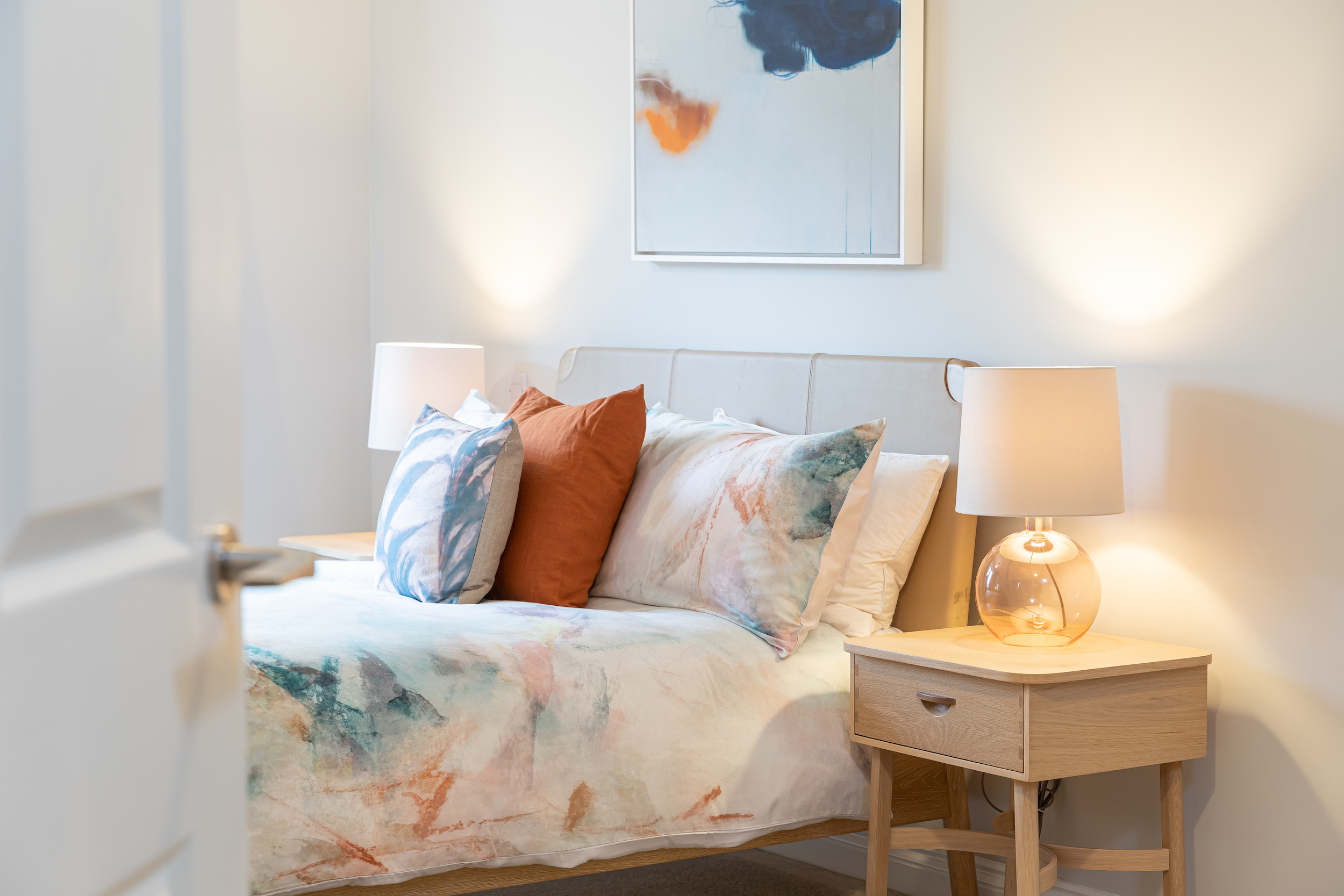 Cozy bedroom with a bed featuring abstract watercolor-patterned bedding and pillows, flanked by wooden nightstands with lit lamps and abstract wall art.
