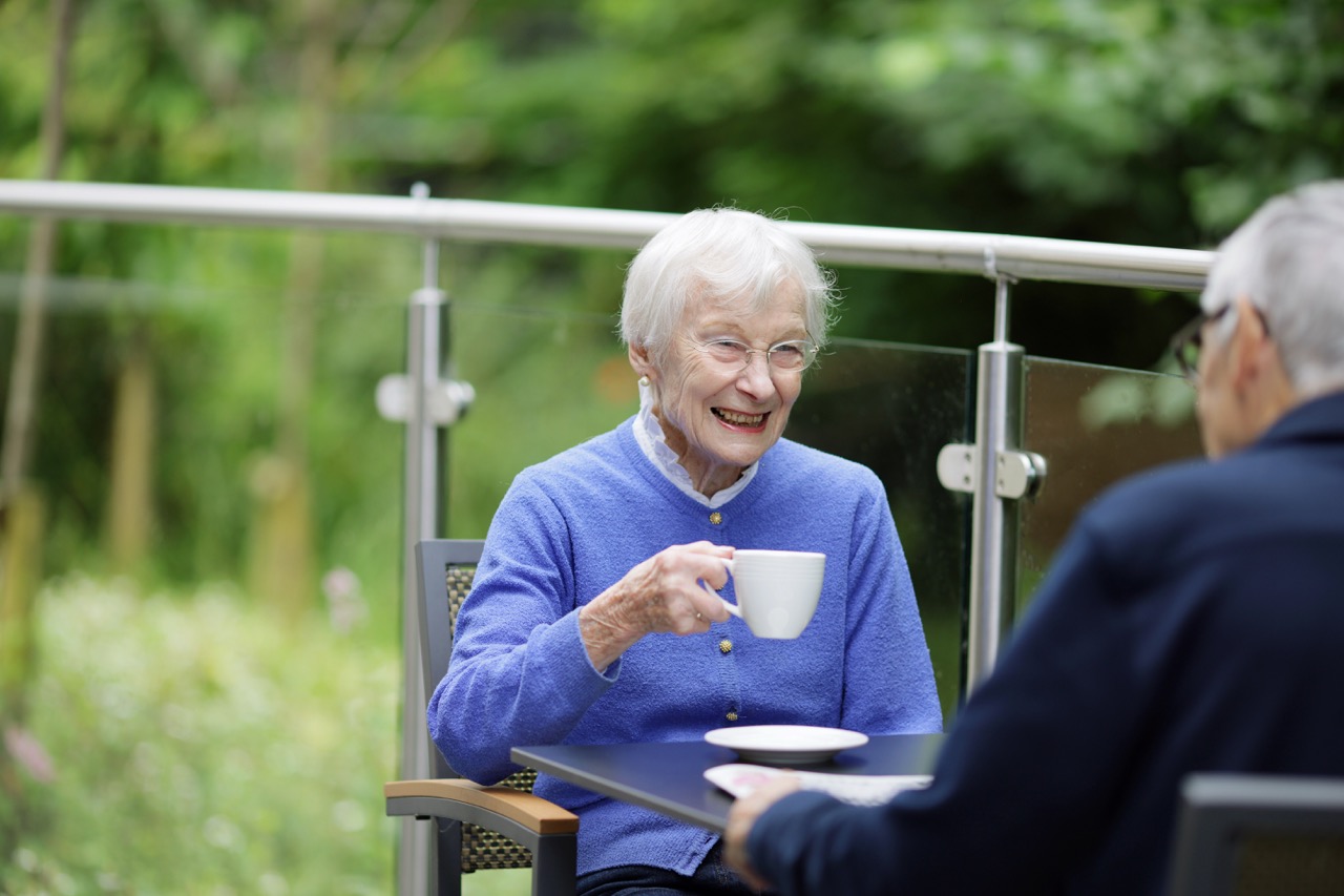 Elderly woman in a blue sweater smiling and holding a white cup while sitting outside at a table with another person.