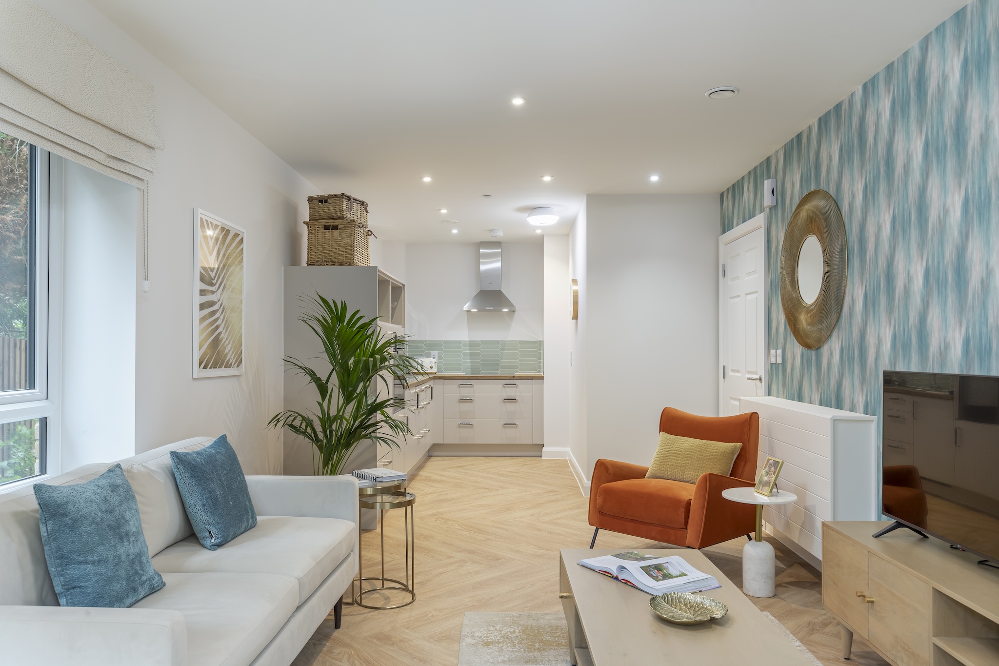 Modern living room with a white sofa and blue pillows, an orange armchair, a wooden coffee table with an open book, and an open kitchen in the background.