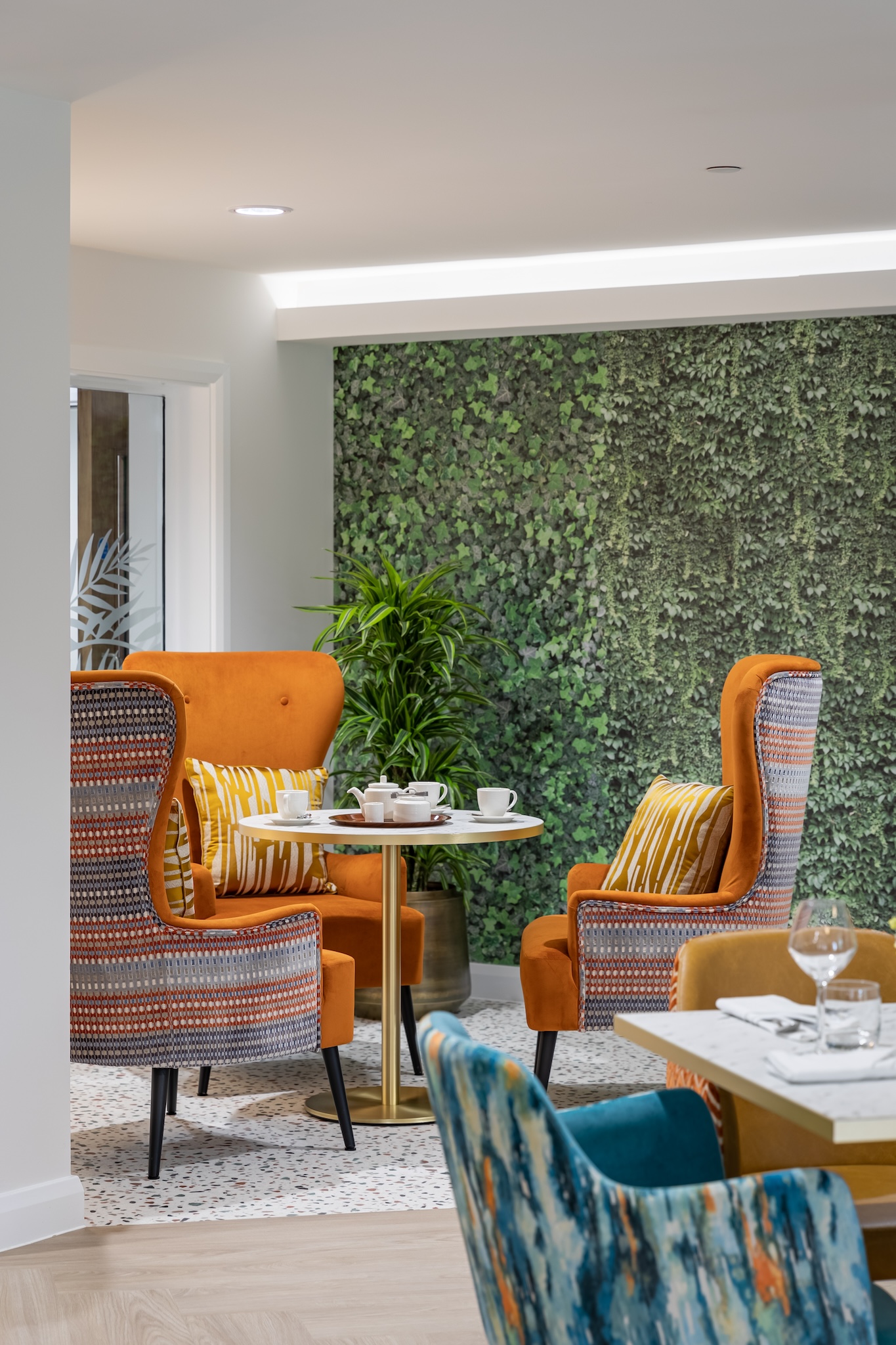 Modern seating area with orange armchairs, a round white table with teacups, a green leafy wall, and colorful patterned chairs in the foreground.