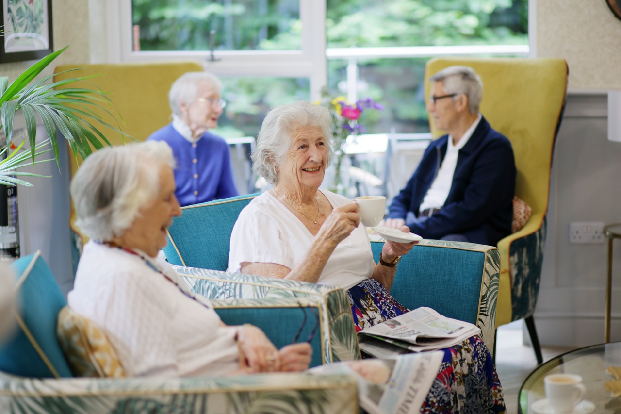 Four elderly women sitting and talking in a bright room, one smiling and holding a cup of tea.