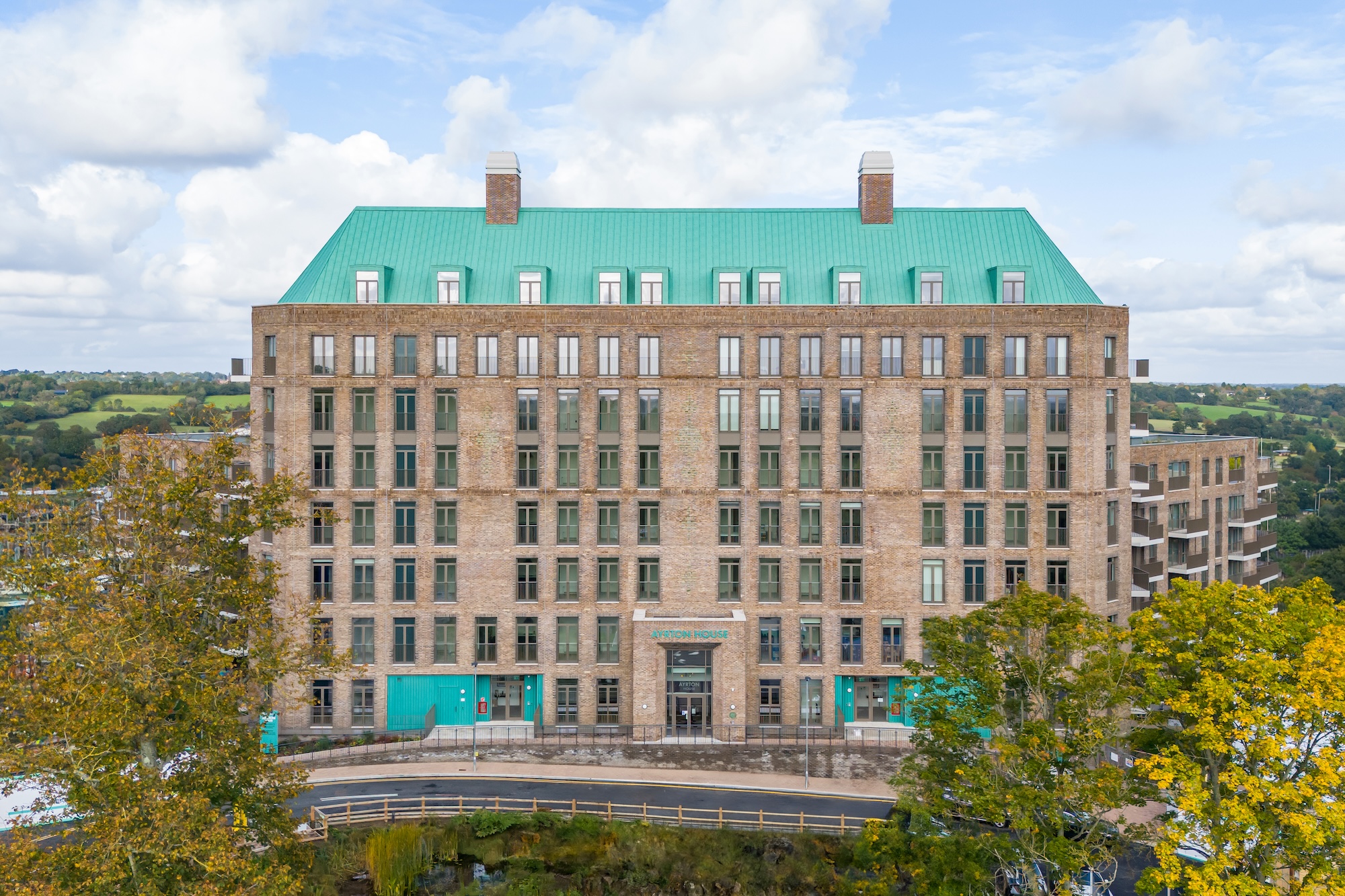 Large brick building with a teal roof labeled Ayrton House, surrounded by trees and countryside.