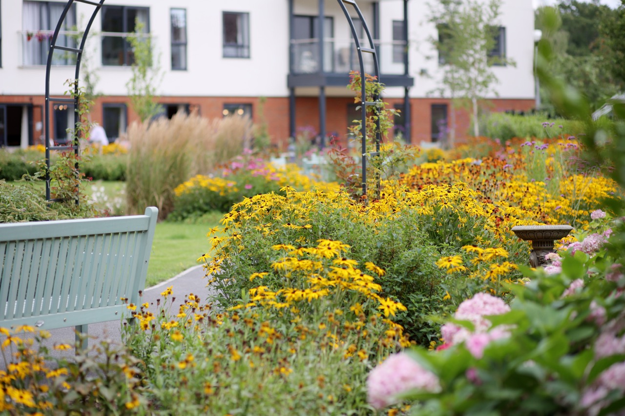 Garden with yellow and pink flowers, a light blue bench, and a metal arch in front of a modern residential building.