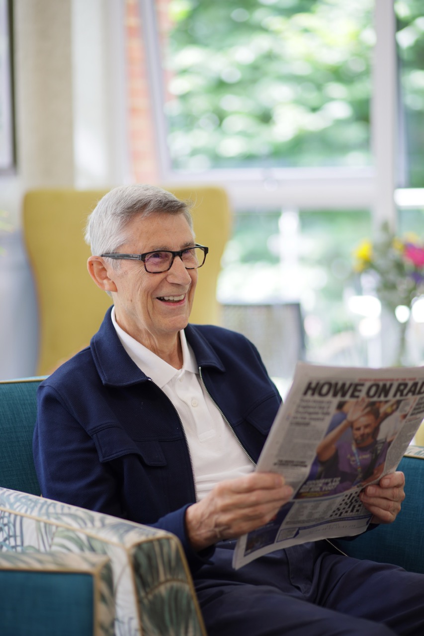 Elderly man with glasses sitting on a blue couch reading a newspaper and smiling.