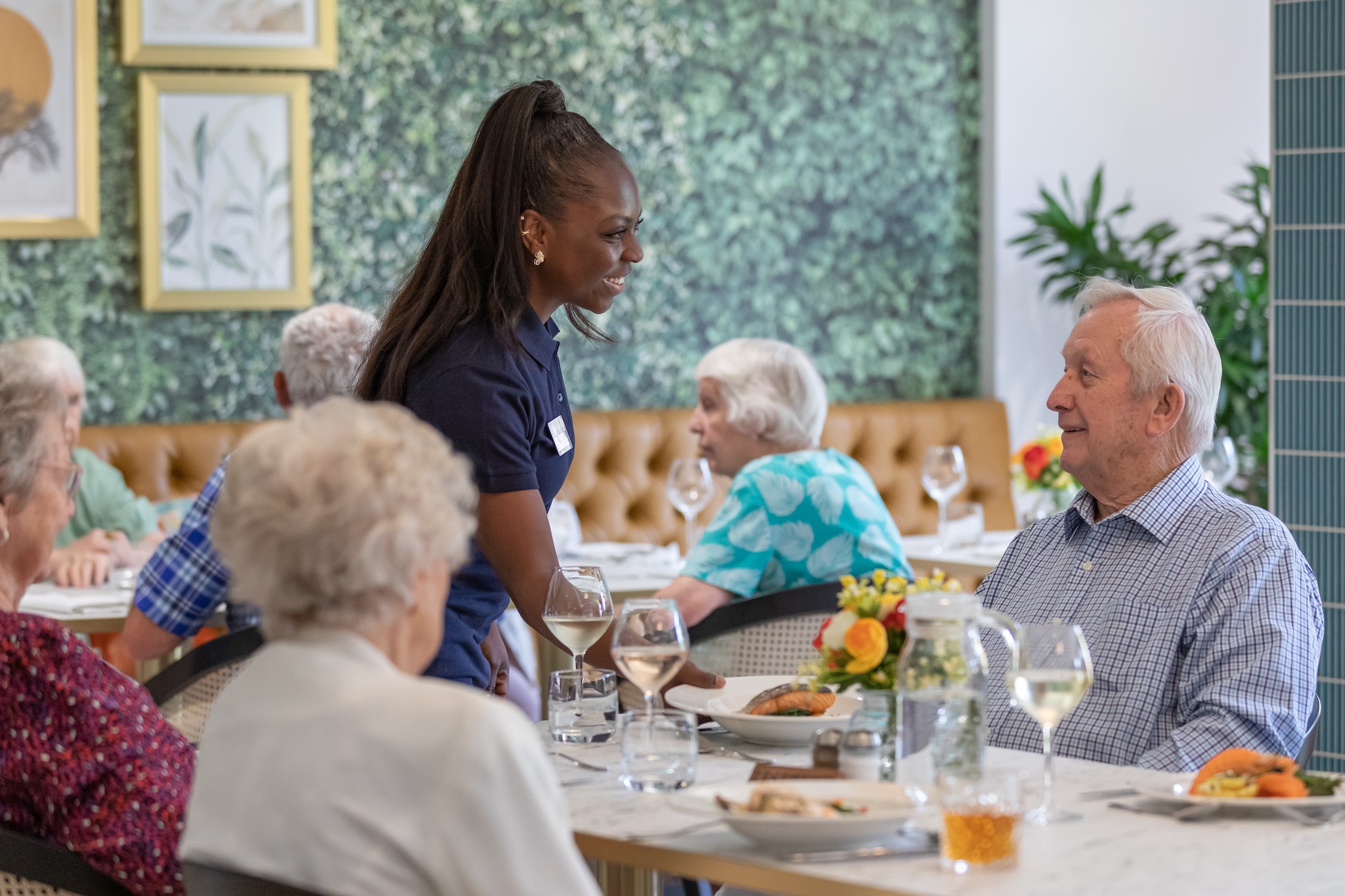 Caregiver smiling and serving food to elderly man seated at a dining table in a bright, decorated room.