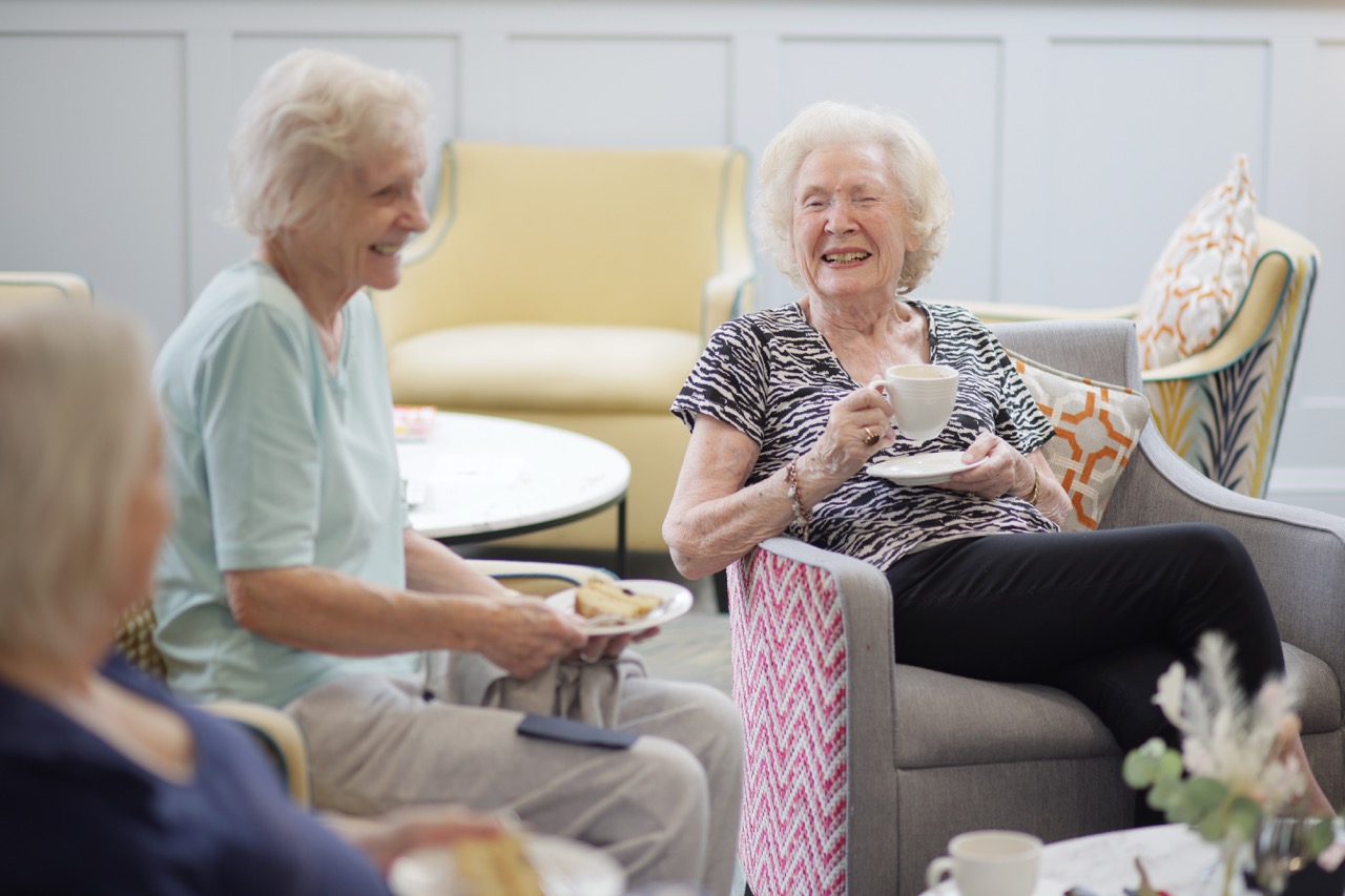 Three elderly women sitting and socializing in a cozy living room, one holding a cup and saucer, another holding a plate with food.