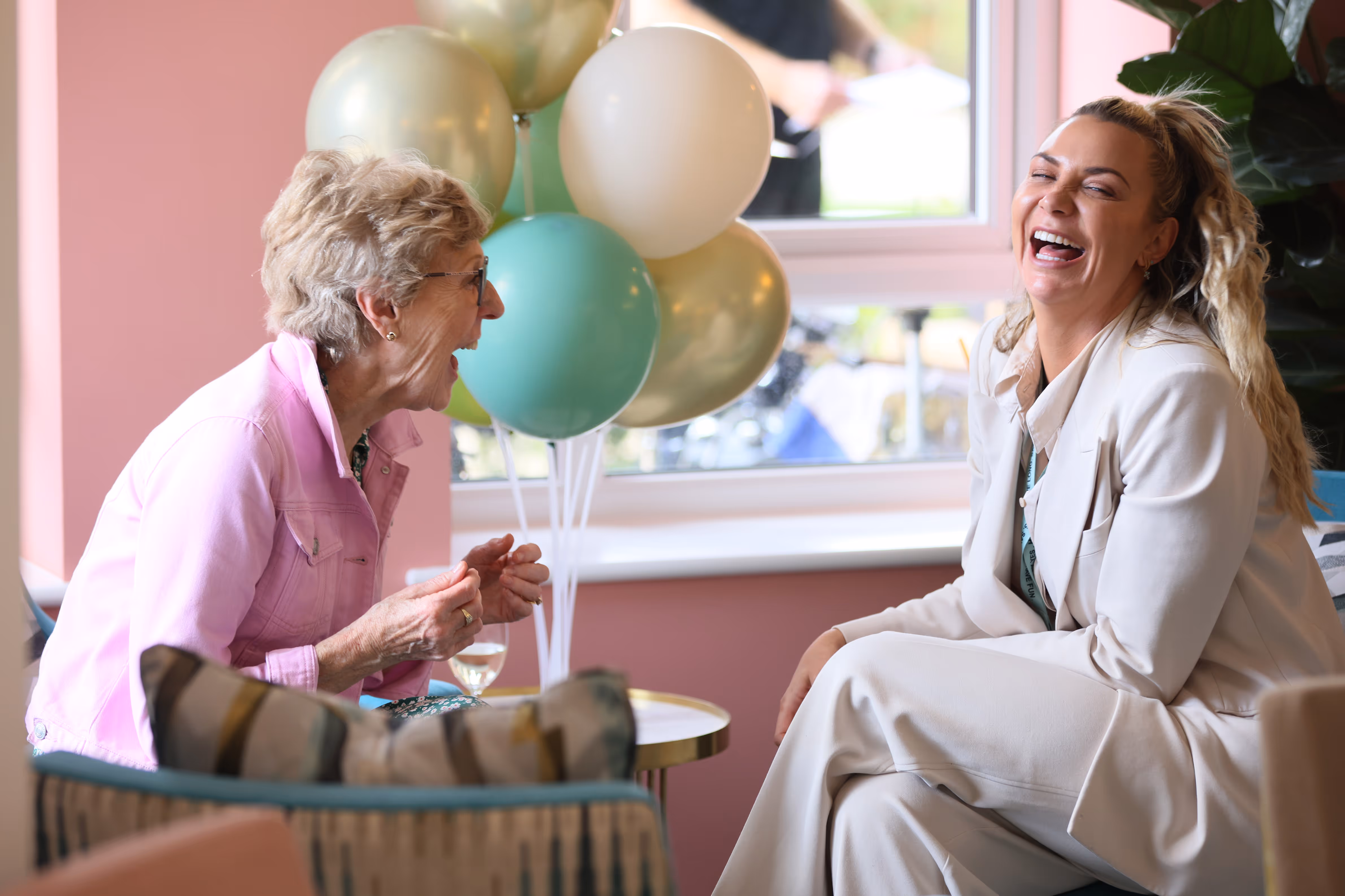 Two women laughing and enjoying a conversation next to a bunch of green and white balloons by a window.