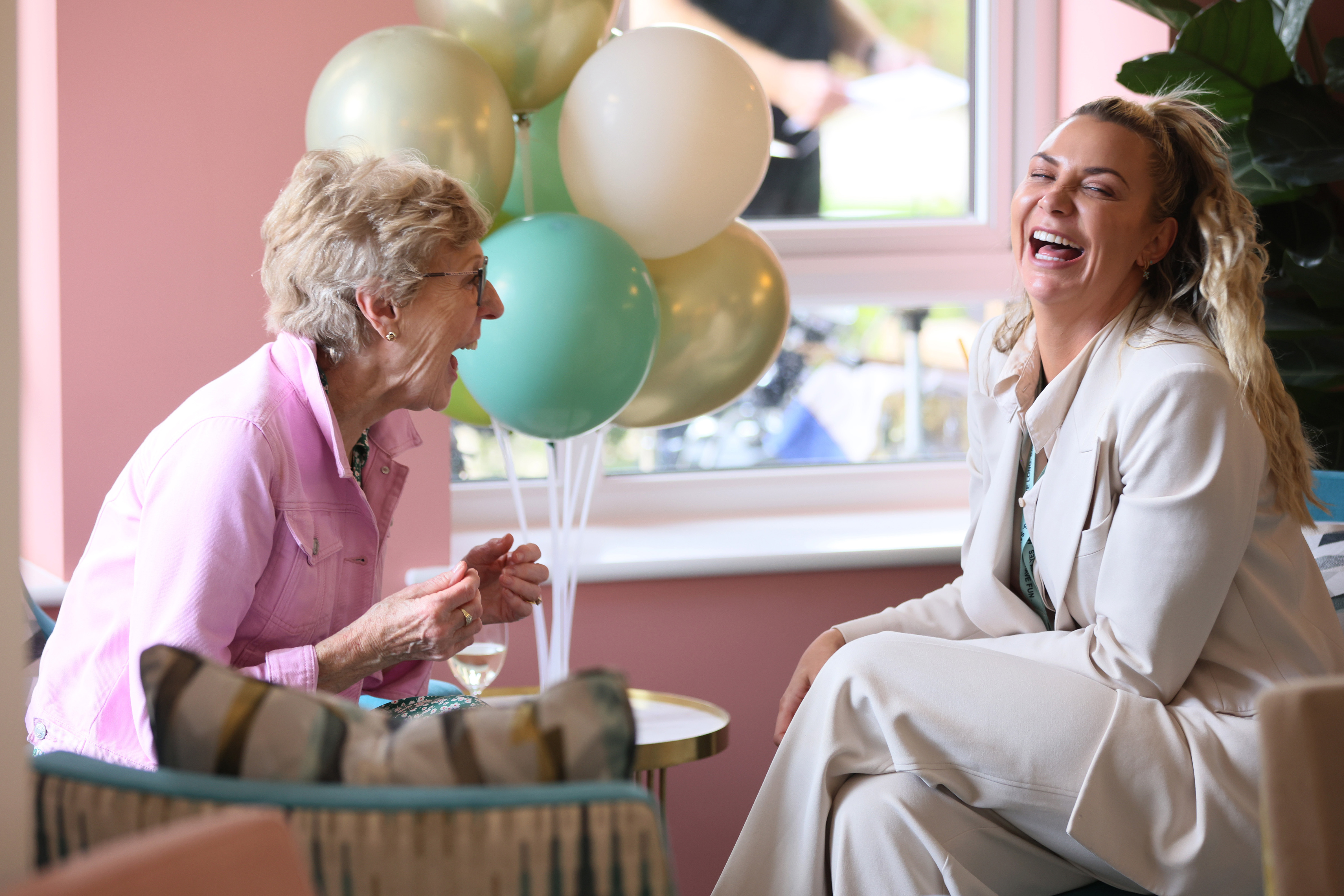 Two women laughing and enjoying a conversation next to a bunch of green and white balloons by a window.