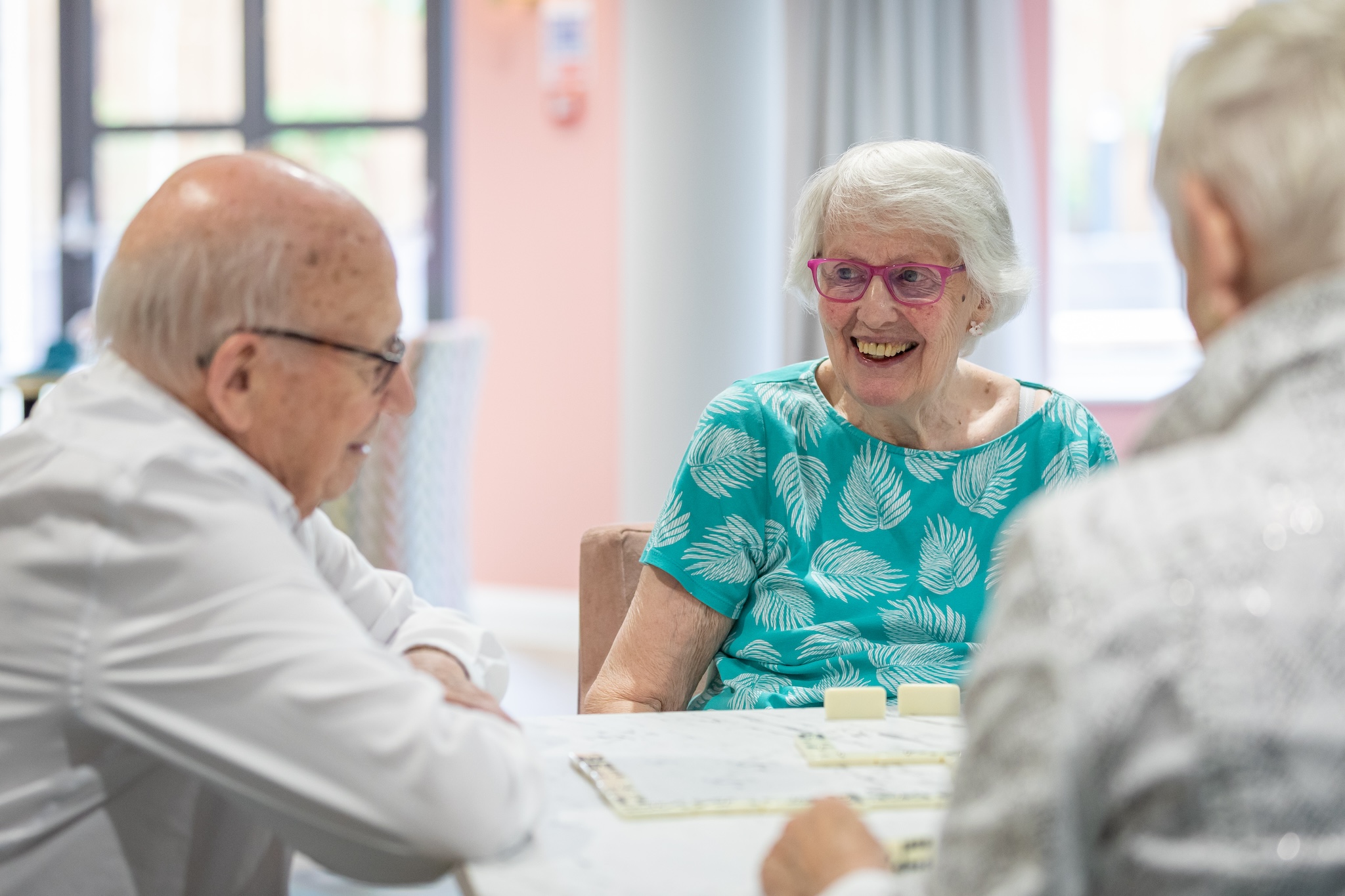 Three elderly people sitting around a table playing a board game, with one woman smiling and wearing pink glasses and a teal shirt with leaf patterns.