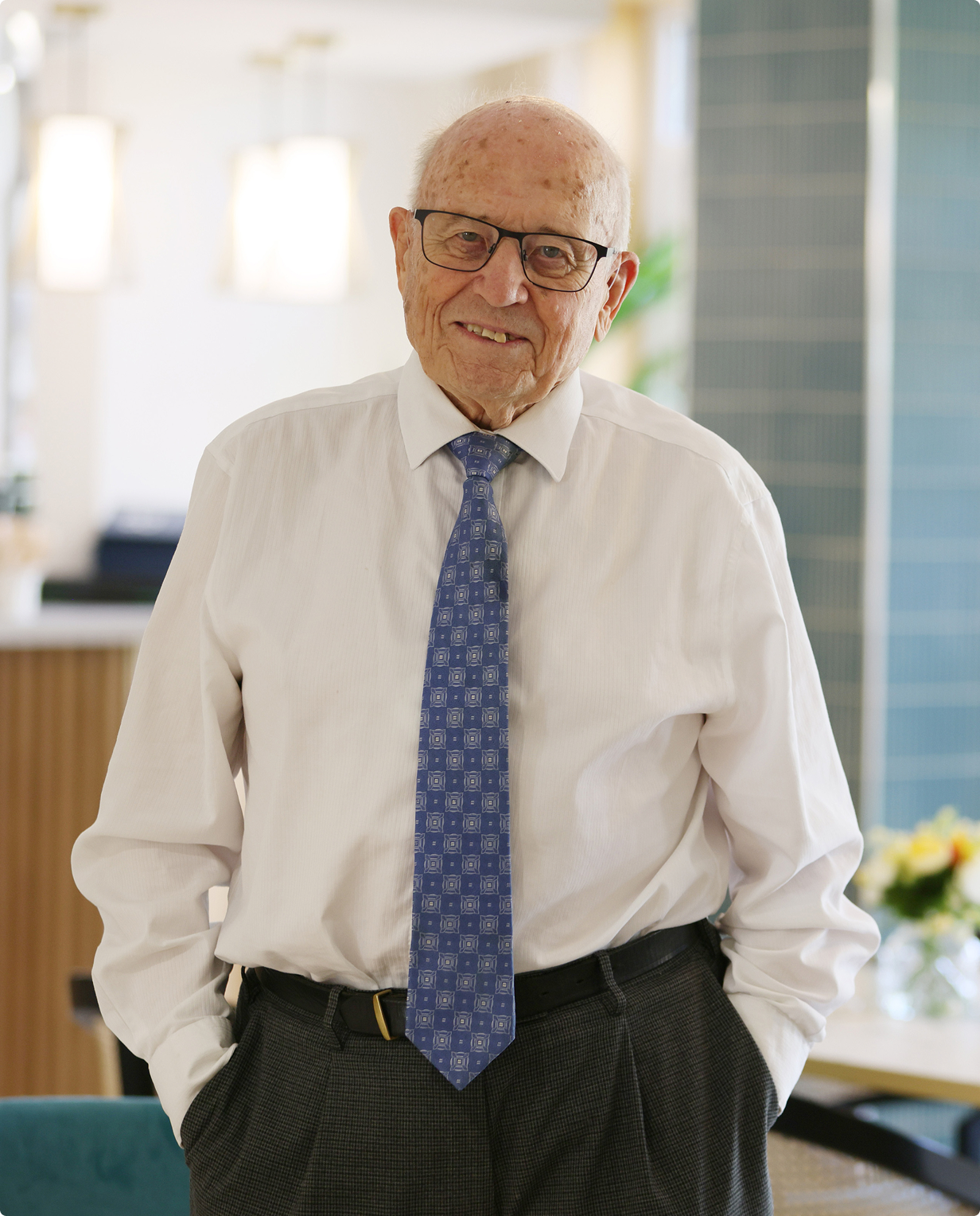 Smiling elderly man wearing glasses, white shirt, and blue patterned tie standing indoors with hands in pockets.