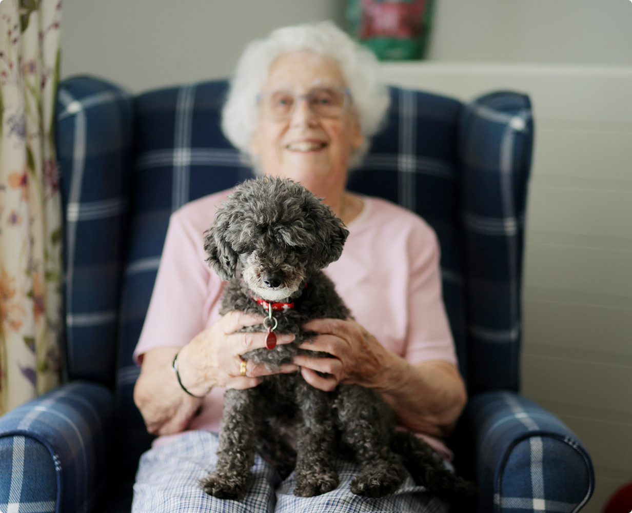 Elderly woman with white hair and glasses sitting on a blue plaid armchair holding a small curly gray dog with a red collar.