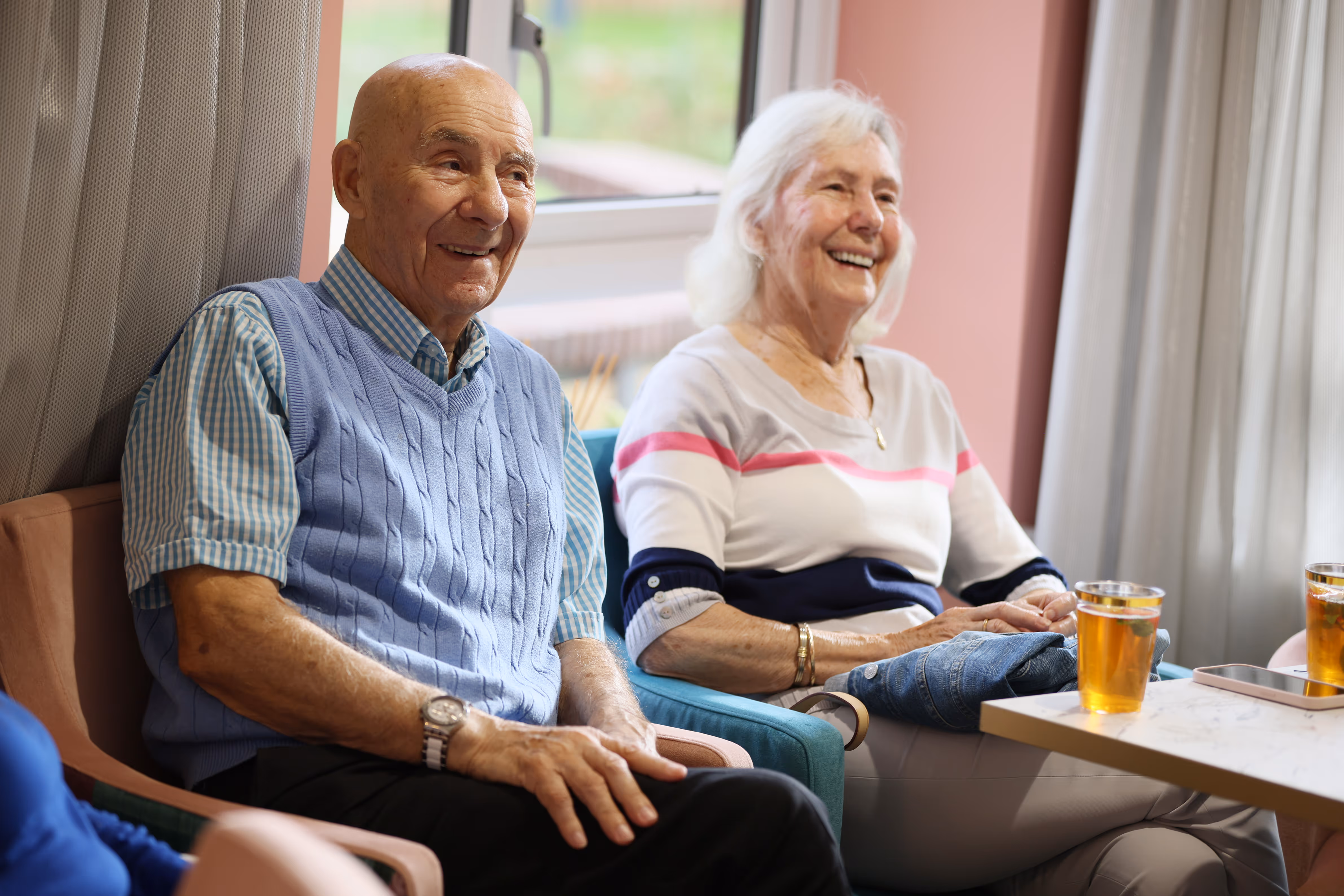 An elderly man and woman sitting indoors, smiling and enjoying a conversation with drinks on the table.