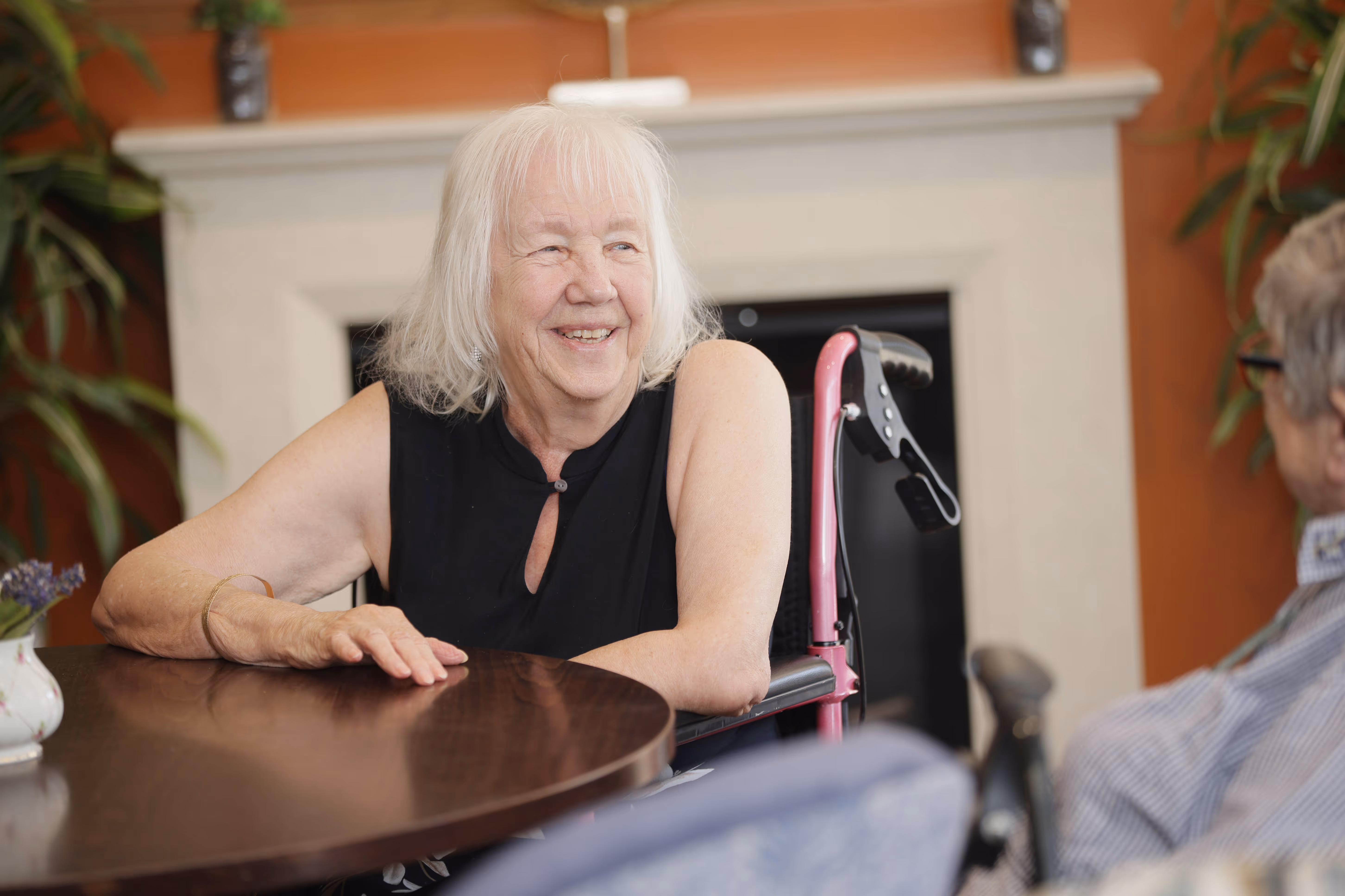 Smiling elderly woman with white hair sitting in a pink wheelchair at a wooden table, talking to a person seated nearby.