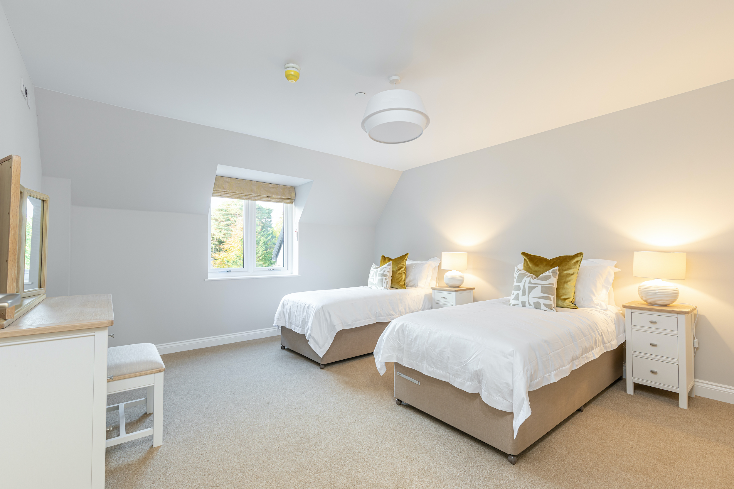 Bright bedroom with two single beds dressed in white linens, beige bed bases, green and patterned pillows, nightstands with lit lamps, a window with a beige blind, and a white dresser with a stool.