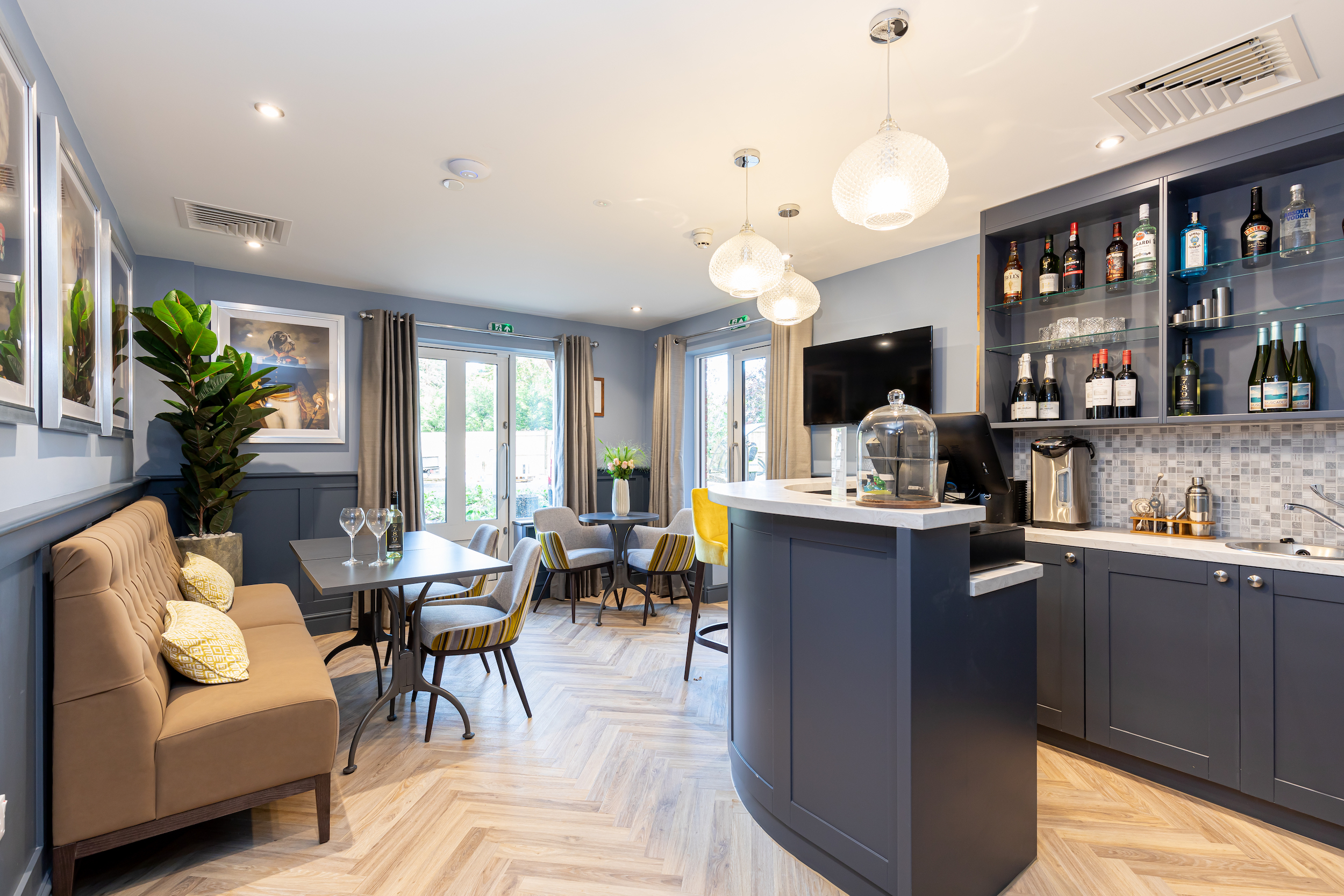 Modern lounge area with gray and beige seating, a curved bar counter stocked with bottles, and large windows with curtains.