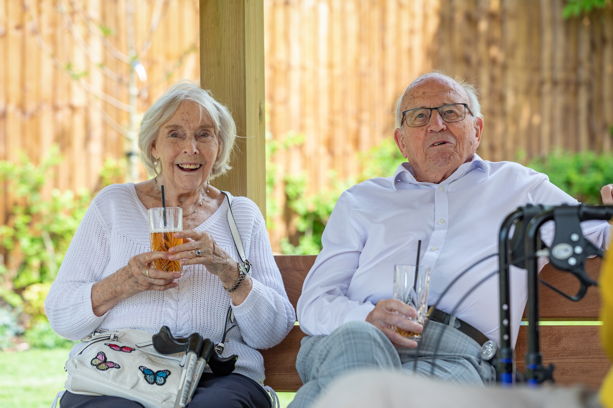 Smiling elderly woman and man sitting outdoors on a bench, each holding a glass with a drink and straws, with walking aids nearby.