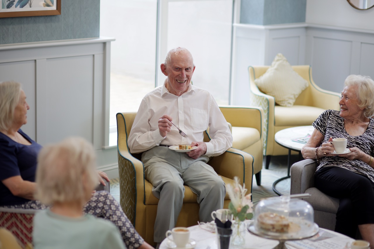 Group of elderly people smiling and socializing in a cozy living room with cups and cake on the table.