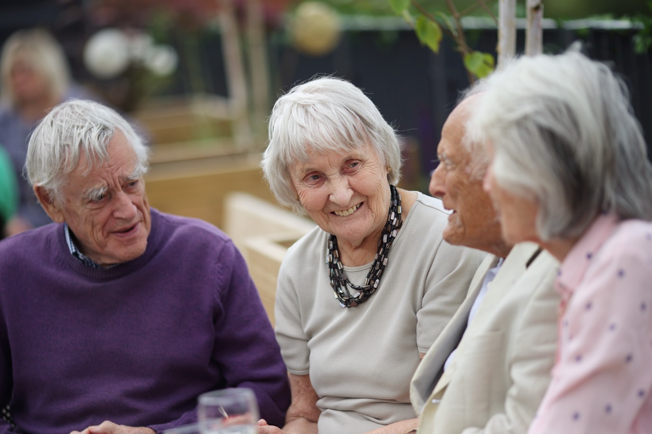 Four elderly people sitting outdoors engaged in a friendly conversation, with one woman smiling.
