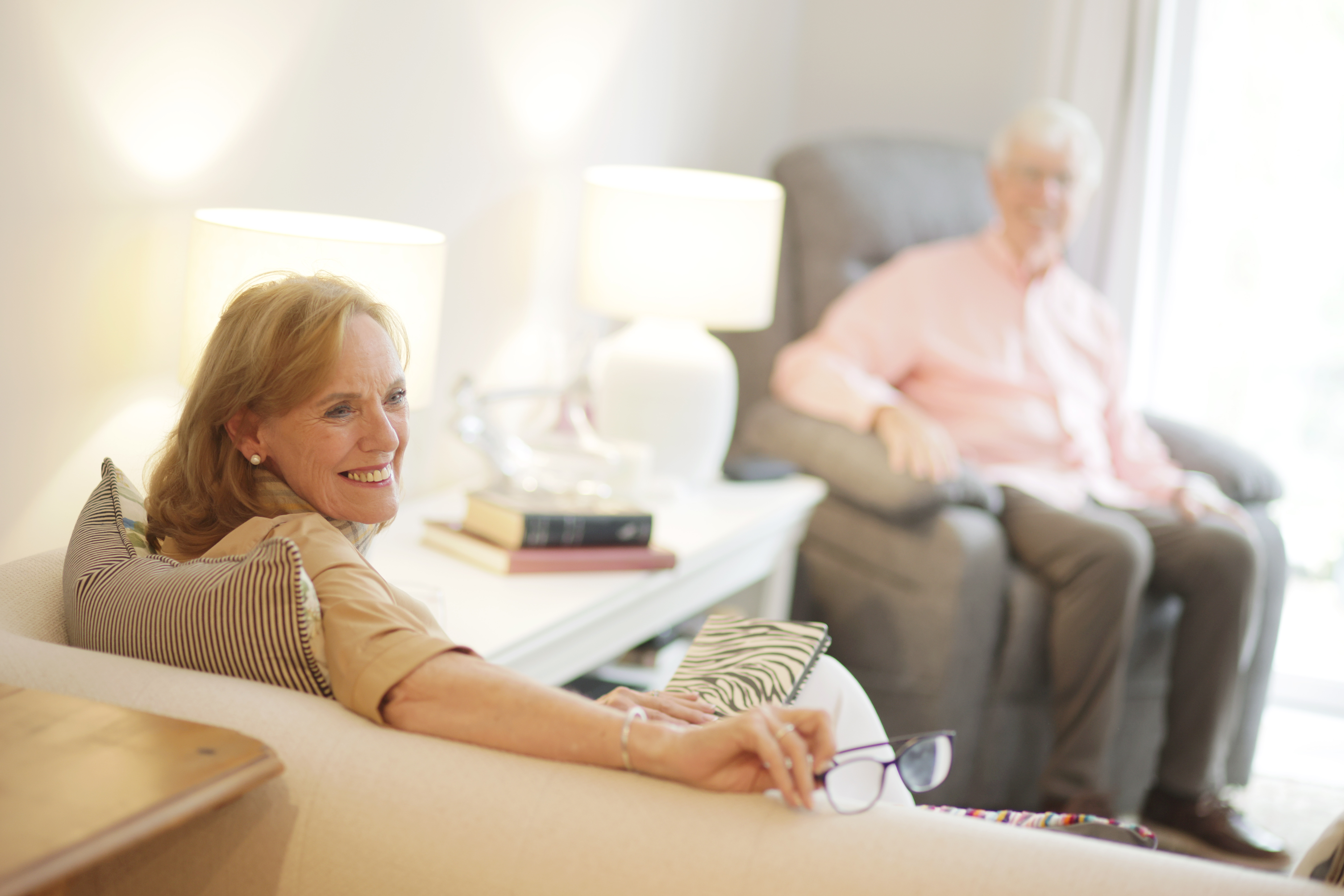 Smiling elderly woman sitting on a beige sofa holding glasses with an elderly man seated in a gray armchair in the background.