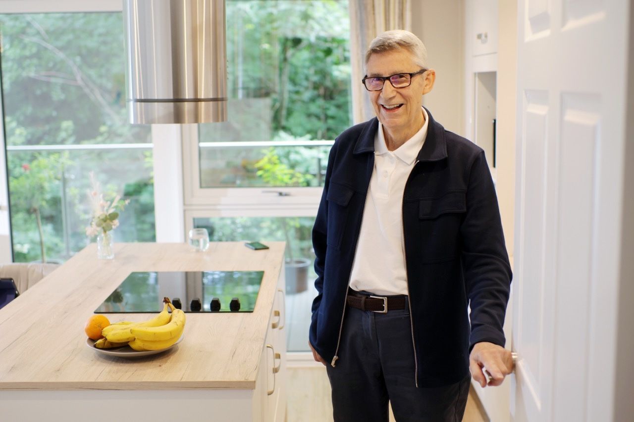 Smiling elderly man wearing glasses and navy jacket standing in a bright kitchen with fruit on the counter.