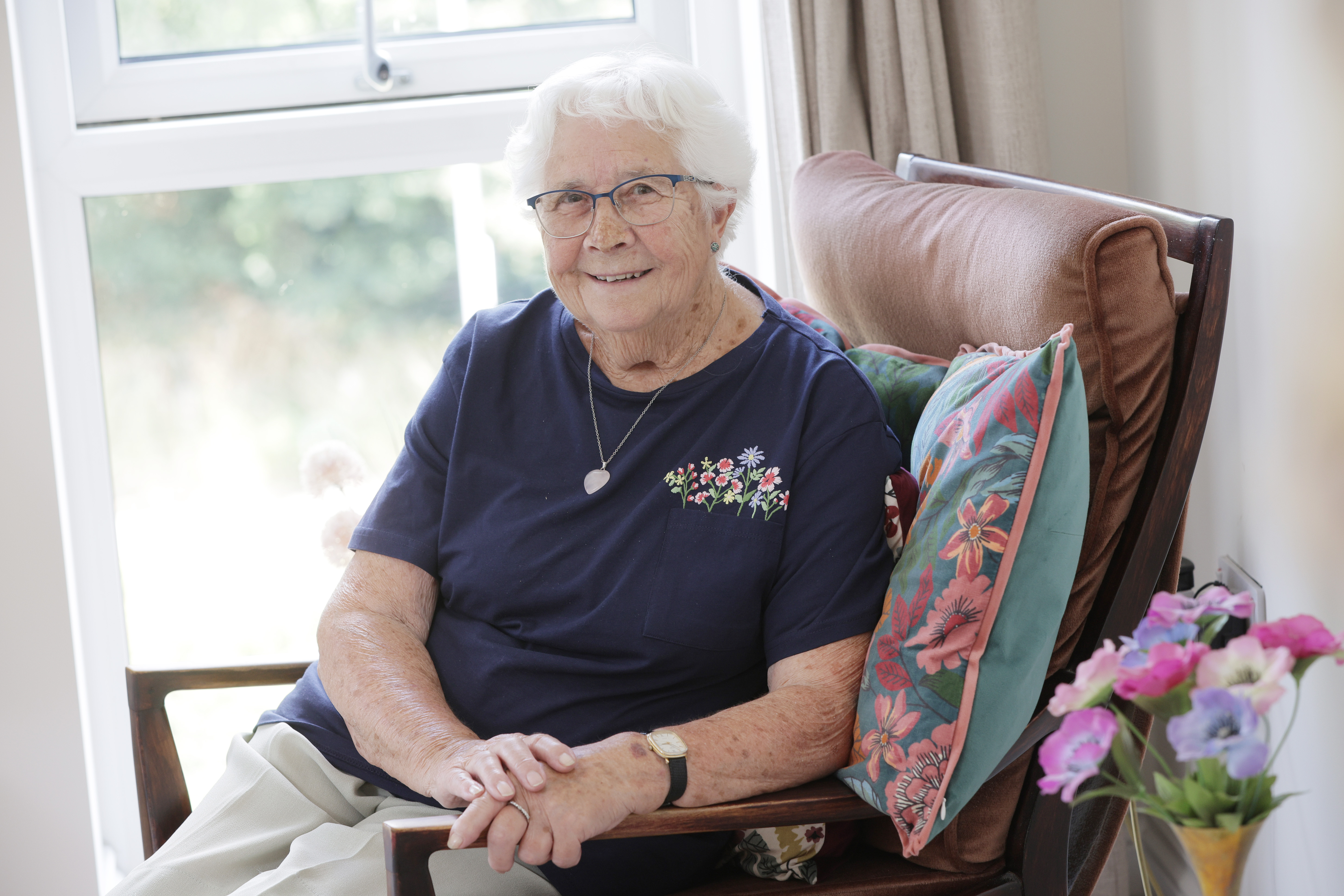 Smiling elderly woman with white hair and glasses sitting in a cushioned chair near a window, wearing a navy shirt with floral embroidery.