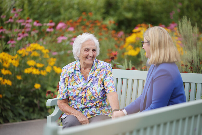Elderly woman smiling and holding hands with a younger woman on a bench in a garden with colorful flowers.