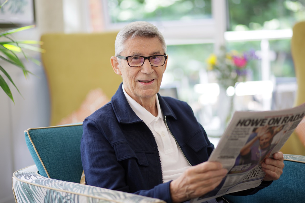 Elderly man wearing glasses and a navy jacket sitting on a patterned chair reading a newspaper indoors.