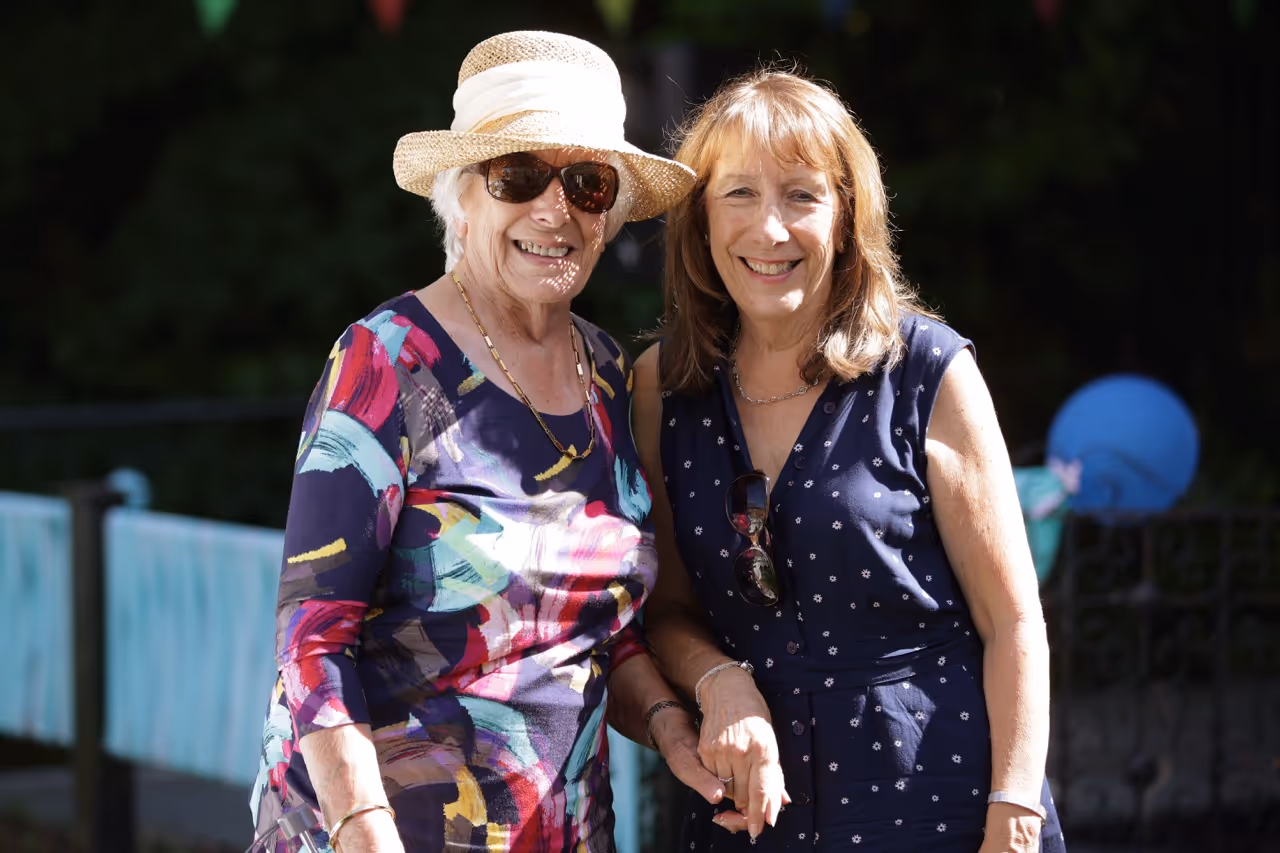Two smiling women standing outdoors, one wearing a colorful dress and straw hat with sunglasses, the other in a navy dress with small white patterns.