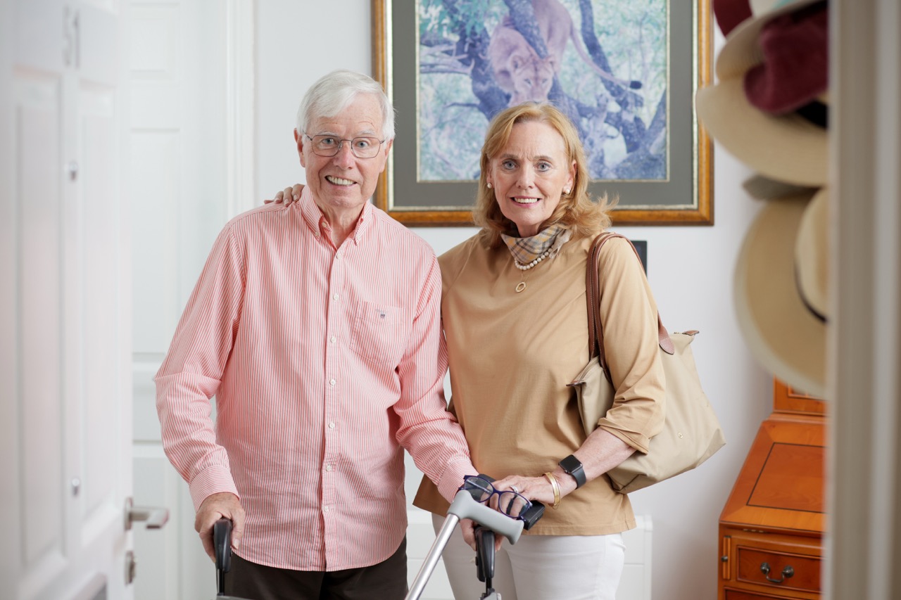 Smiling elderly man using a walker standing next to a woman with her hand on his shoulder in a well-lit room with framed artwork and hats on a wall.