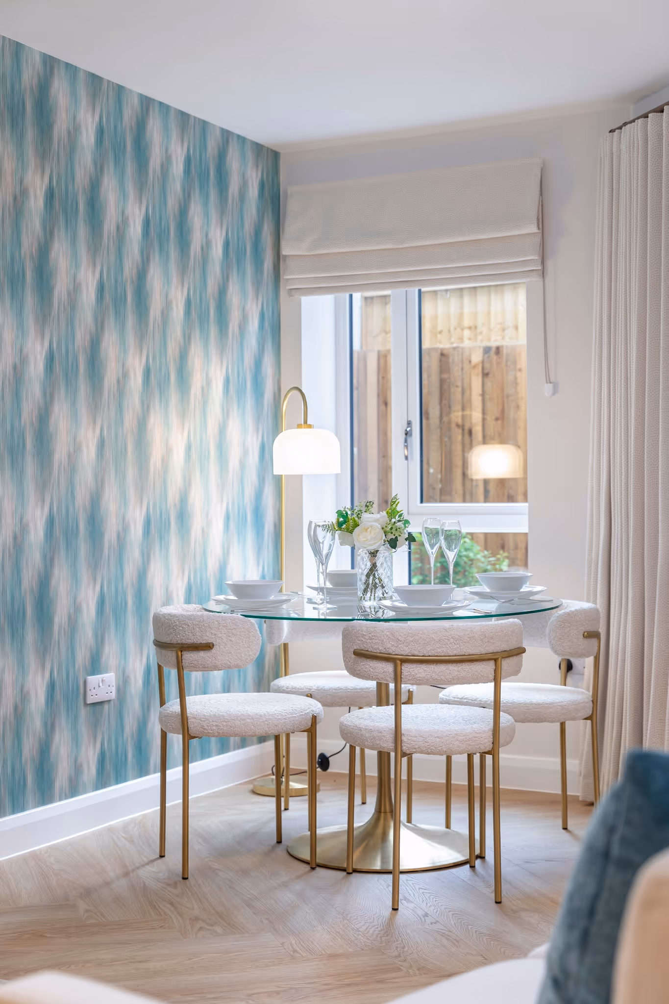 Modern dining area with a round glass table, four white upholstered chairs with gold legs, a floral centerpiece, and a floor lamp next to a window with beige curtains.