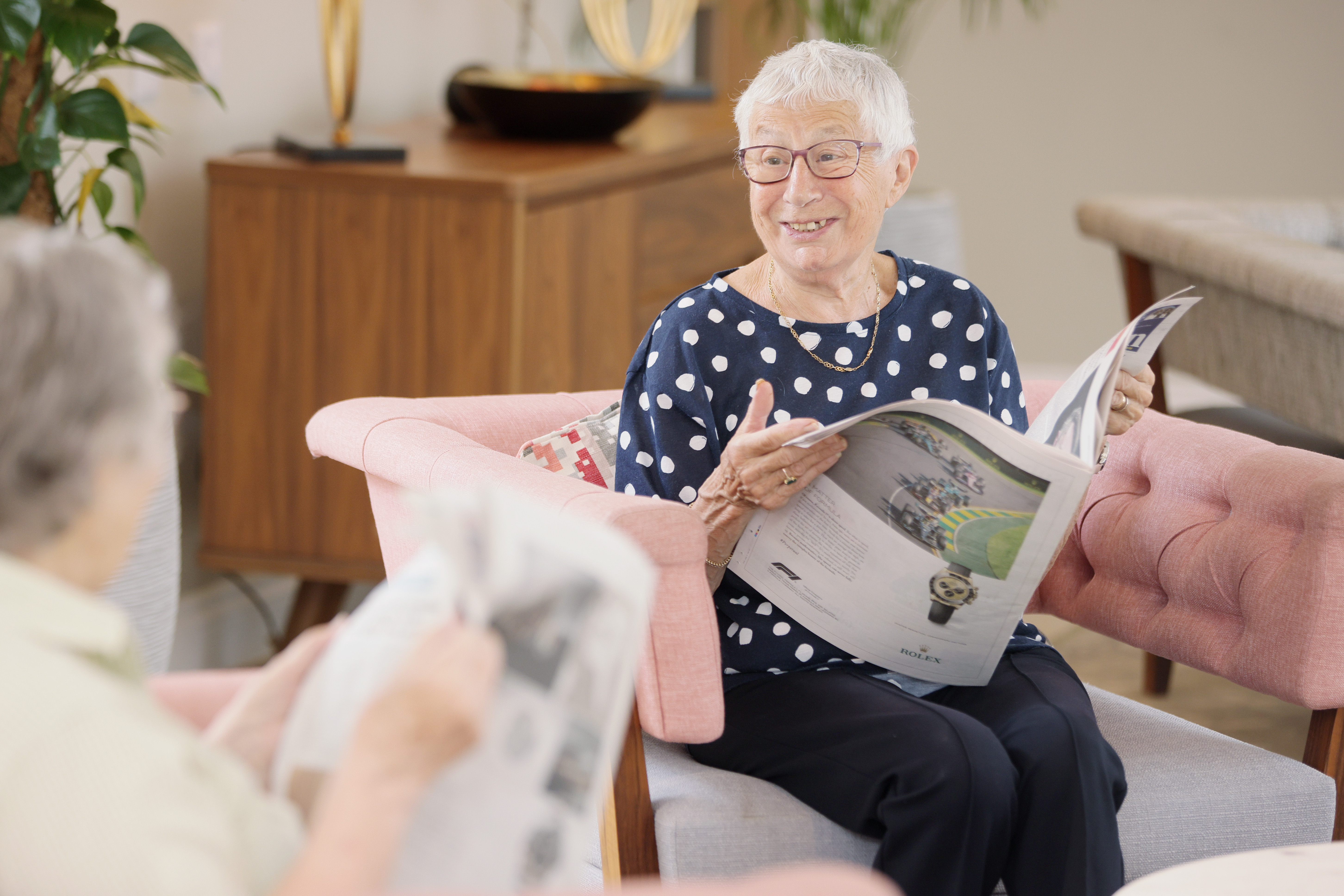 Smiling elderly woman with short white hair and glasses sitting on a pink chair, reading a newspaper and looking at another person.