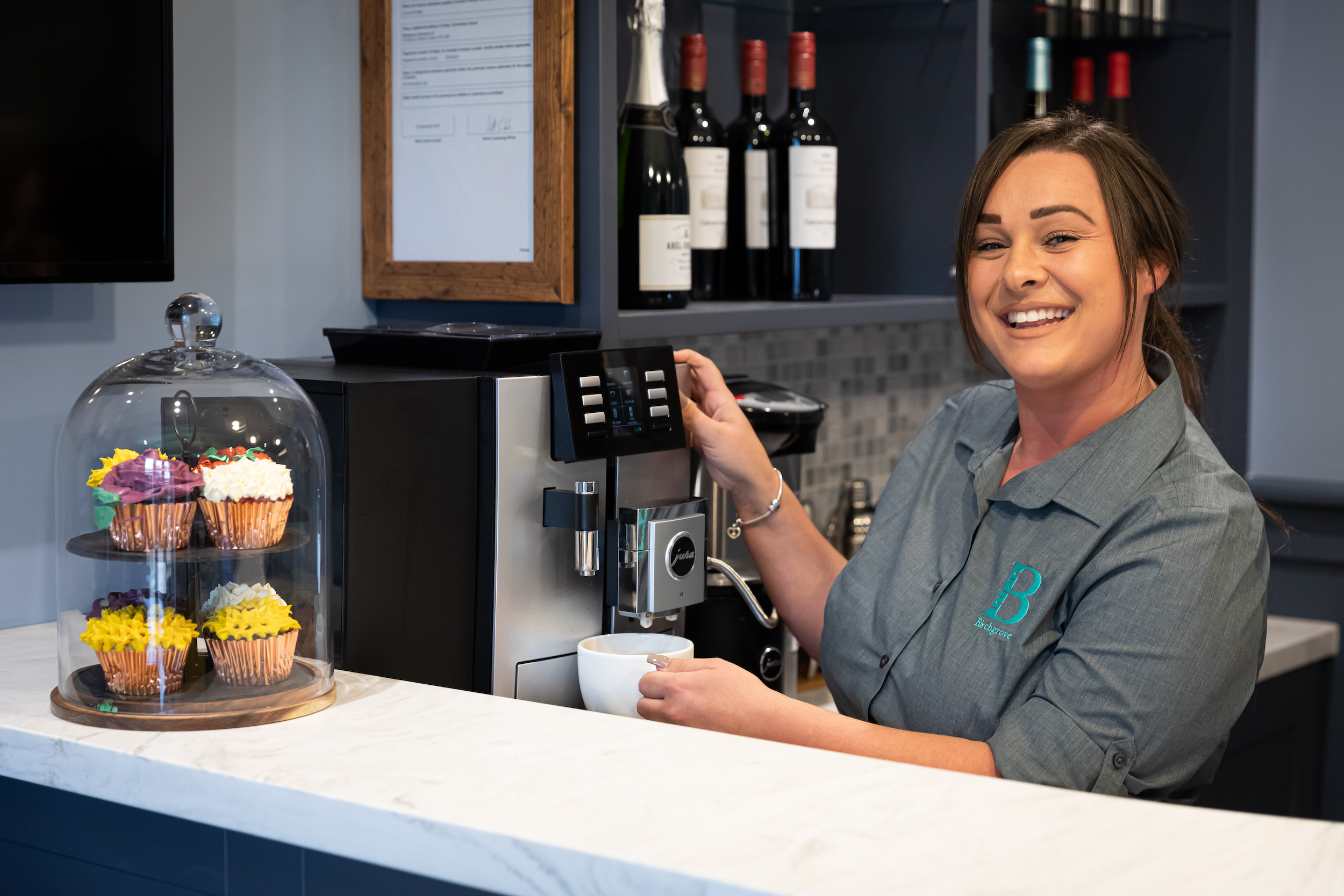 Smiling woman operating a coffee machine with a cup, with colorful cupcakes under a glass dome nearby.
