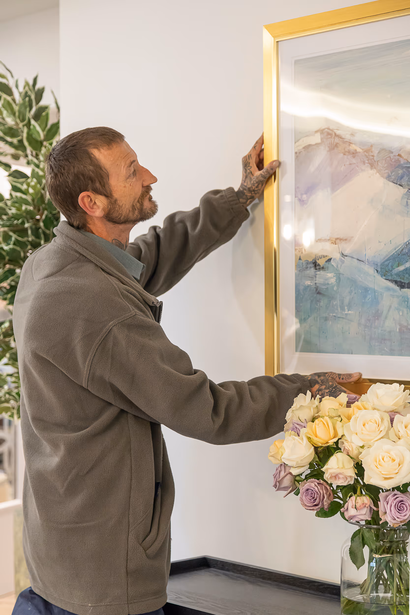 Man with tattoos hanging a framed abstract painting on a white wall next to a bouquet of pastel roses in a glass vase.
