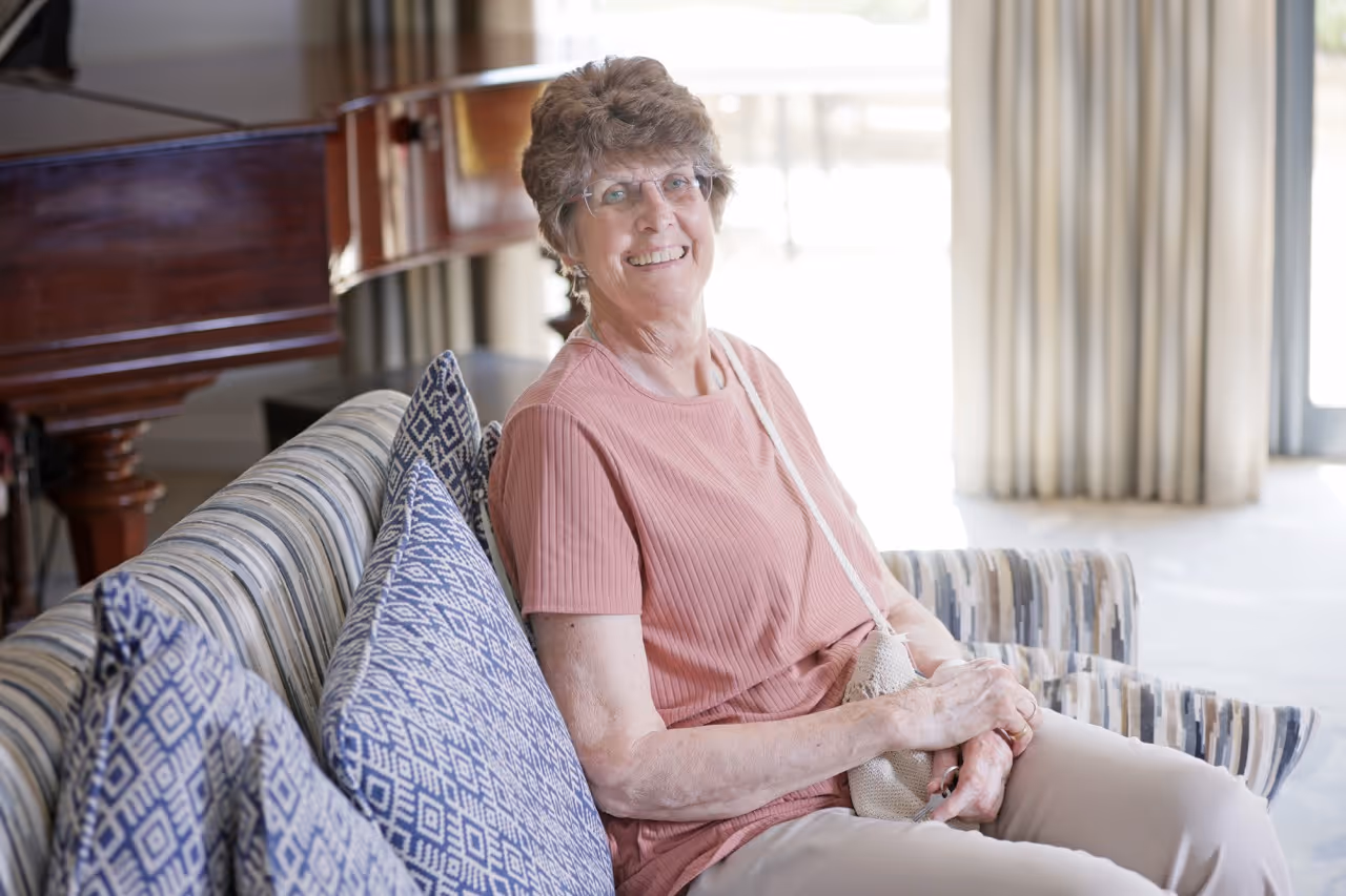 Smiling elderly woman wearing glasses sitting on a patterned sofa in a well-lit room.